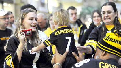 Brittany Jenkins, of Cabot,  reaches for her son, Caleb, as former Steeler L.C. Greenwood passes him back after a photo yesterday during the Steelers tailgate party at The Galleria at Pittsburgh Mills in Frazer.