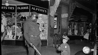 Small boy in police officer's uniform with Prince "Big Blue" Bruce on beat outside Alpern's clothing store with beauty parlor next door, c. 1952.