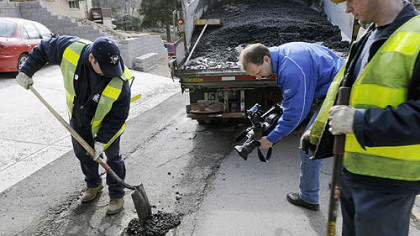 Potholes were the center of media attention on Carnahan Road in Banksville yesterday. Public works employee Mike Maiorano is photographed for television news while filling one hole while fellow employee Eric Thomas waits to tamp the material down.
