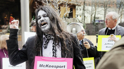 Ankur Mehta, who has lived in Braddock for two years, plays a zombie as he joined about 30 people protesting the closing of UPMC Braddock Hospital. The group, including filmmaker Tony Buba, center, of Braddock Hills and David Hughes, who plays the part of UPMC President and CEO Jeffrey Romoff, did their theater in front of the UPMC headquarters Downtown.